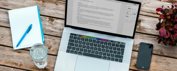Ordinateur portable ouvert sur un résumé exécutif, avec un carnet, un stylo, un verre d'eau et un smartphone sur une table en bois.